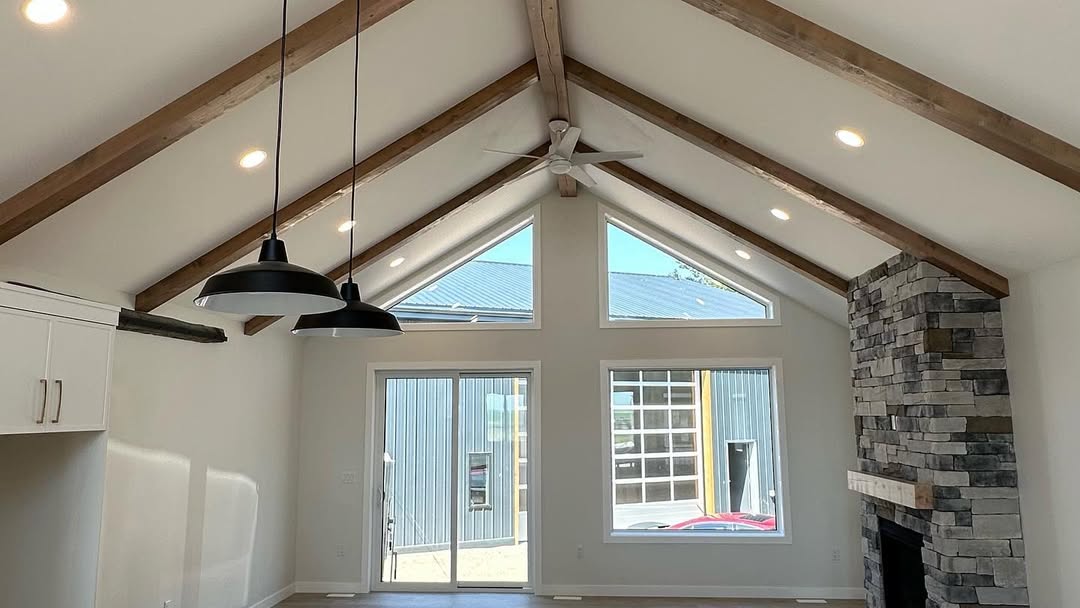 A spacious living room with vaulted ceilings and wooden beams, featuring large windows, a stone fireplace, and modern pendant lights, creating an airy and inviting atmosphere.