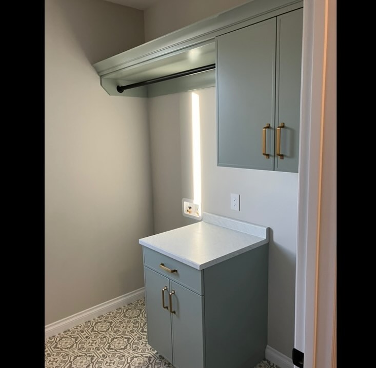Compact laundry room with light green cabinets, gold handles, and a white countertop. A hanging rod is above, and patterned floor tiles add elegance.