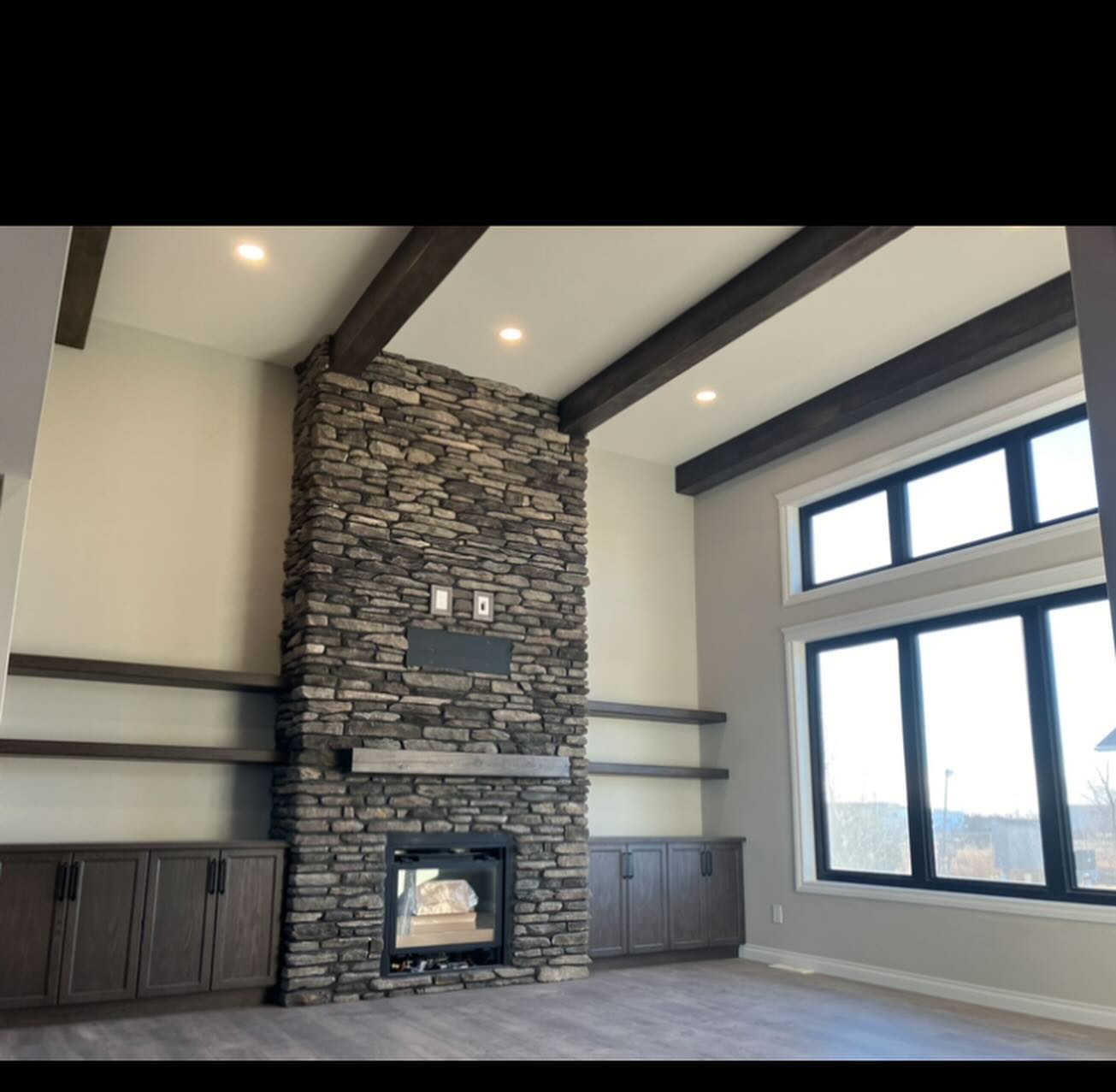 Spacious living room with a tall stone fireplace and wooden mantel. Dark wood cabinetry and shelves flank the fireplace. Large windows provide natural light.