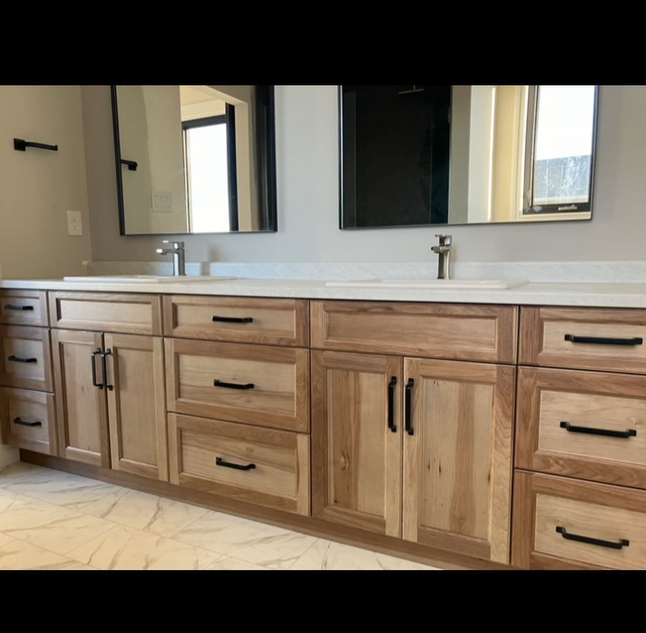 A modern bathroom features dual sinks with sleek silver faucets, set atop a light countertop. Wooden cabinets below have black handles, complemented by large mirrors above. The space is clean and minimalistic, evoking a sense of calm and elegance.