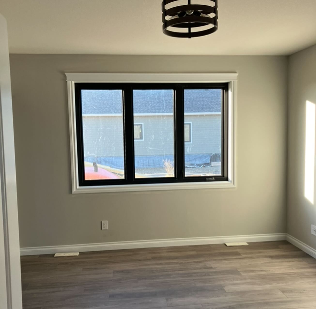 A sunlit, empty room with light gray walls and wood flooring. A large window with black trim offers a view of a building, creating a calm ambiance.