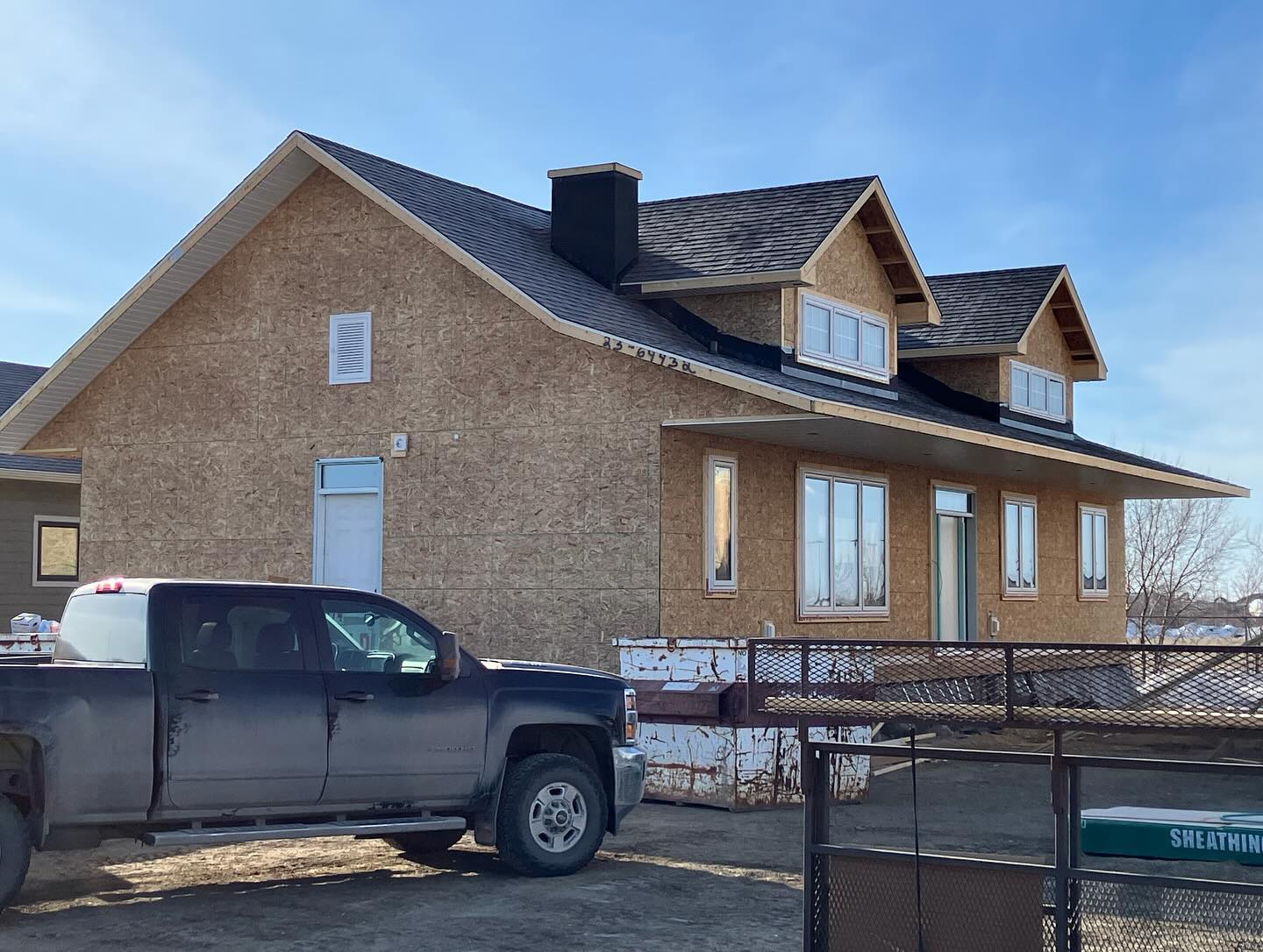 A partially constructed house with a dark shingle roof and unfinished exterior walls. A black pickup truck is parked in front, under a clear blue sky.