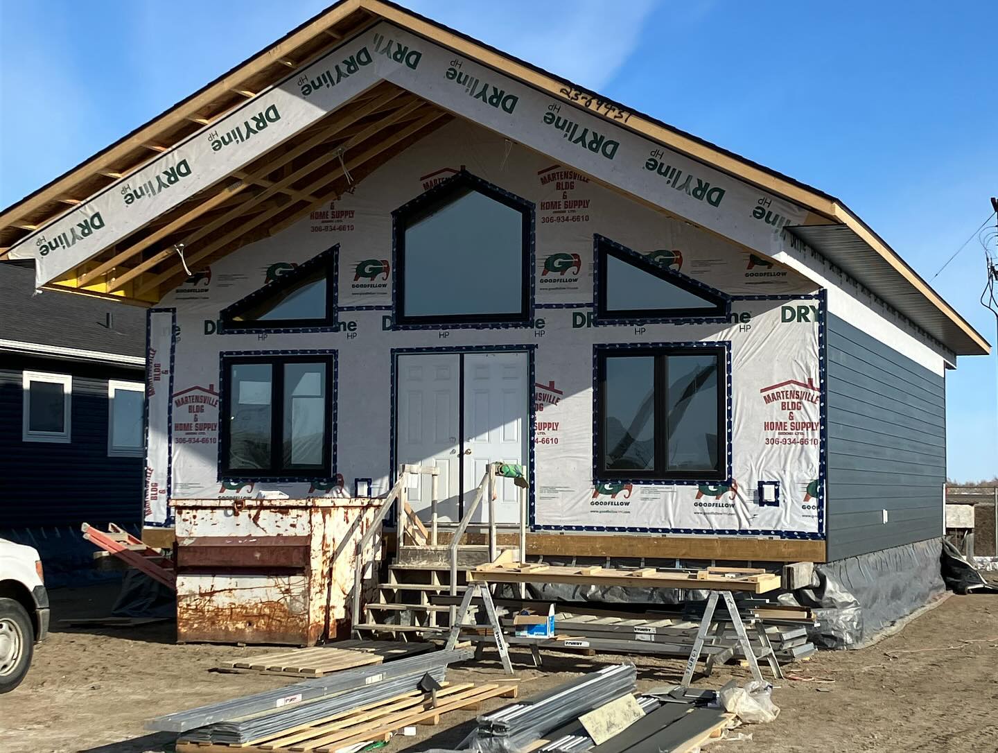 A partially constructed house under clear skies, showcasing exposed wooden beams and partially installed windows. Construction materials are scattered in the foreground.