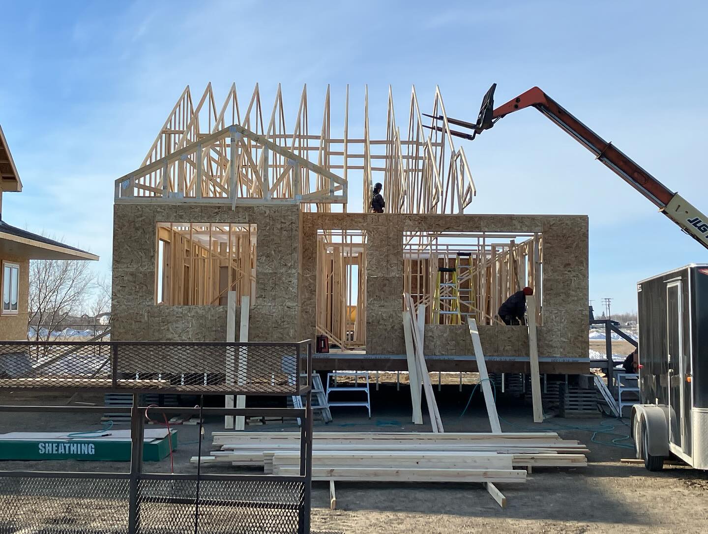 A partially built house in progress with exposed wooden framing and sheathing. Workers and a crane are visible under a clear blue sky, conveying industriousness.