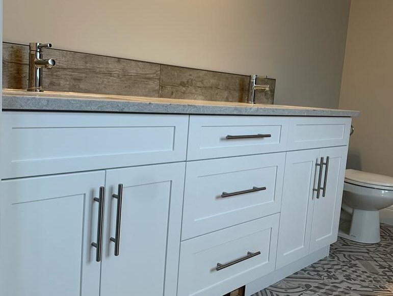 Modern bathroom vanity with dual sinks, sleek white cabinets, silver handles, and a gray countertop. Patterned tile floor and visible toilet create a clean, minimalist look.