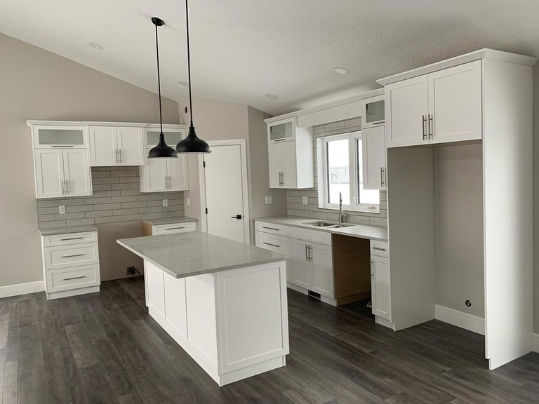 Modern kitchen with white cabinets, stainless steel handles, and a gray tiled backsplash. Features a gray island, dark wood floors, and two black pendant lights.