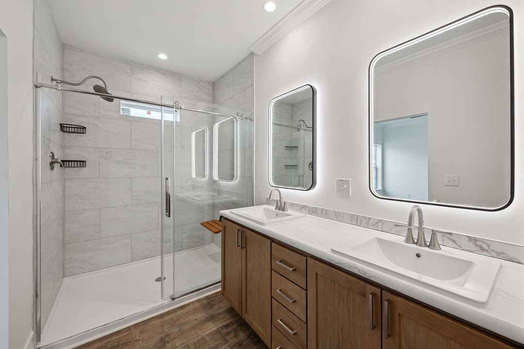 Modern bathroom with dual sinks, illuminated mirrors, and a spacious glass shower with tiled walls. The ambiance is sleek and contemporary.
