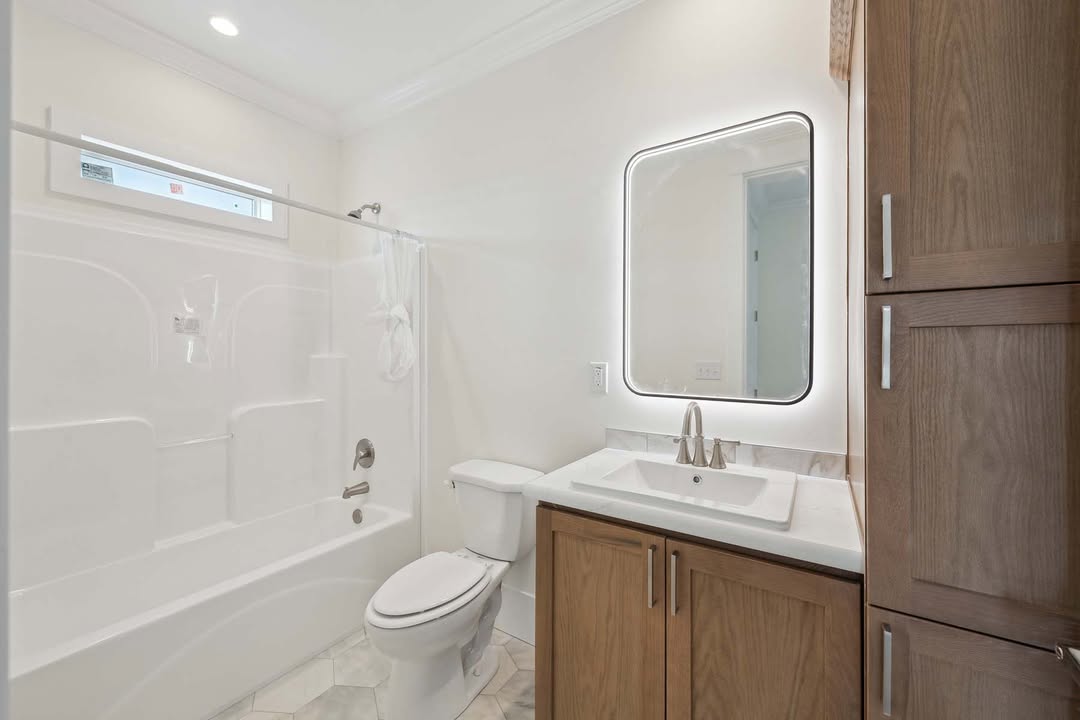Modern bathroom with a white bathtub and curtain, toilet, and a wooden vanity. A backlit mirror adds a touch of elegance and warmth.