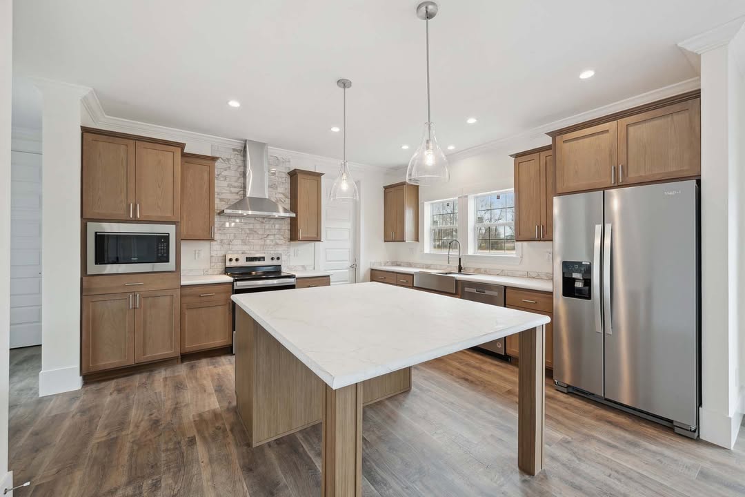Spacious kitchen with wooden cabinets, marble island, pendant lights, and stainless steel appliances, including a fridge and oven. Warm, inviting tone.