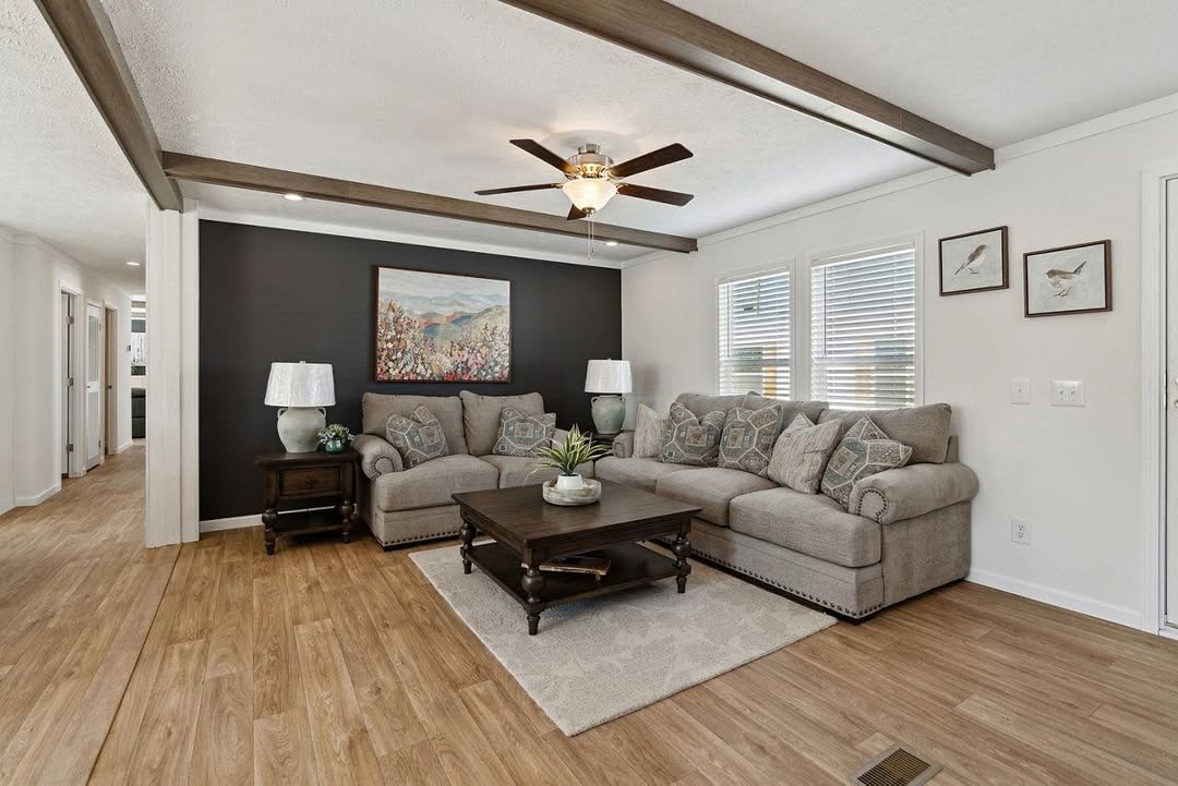 Cozy living room with a large gray sectional sofa, wooden coffee table, and two lamps. Ceiling fan, wood beams, and floral art on a dark accent wall.