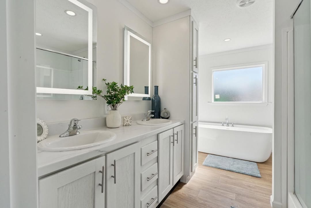 Bright modern bathroom with white cabinets and dual mirrors. Potted plant and decor on counter. Freestanding tub by fogged window. Wood floor adds warmth.