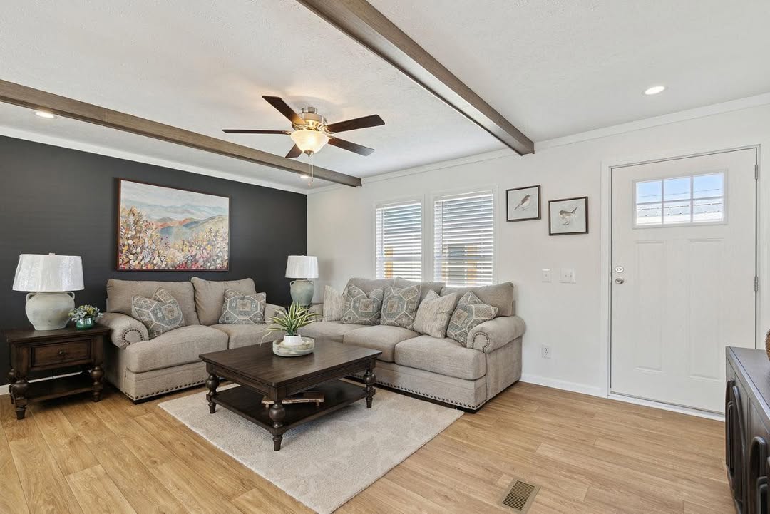 Cozy living room with beige sectional sofa, dark wood coffee table, and wall art. Black accent wall, white lamps, ceiling fan, and wood beams add warmth.