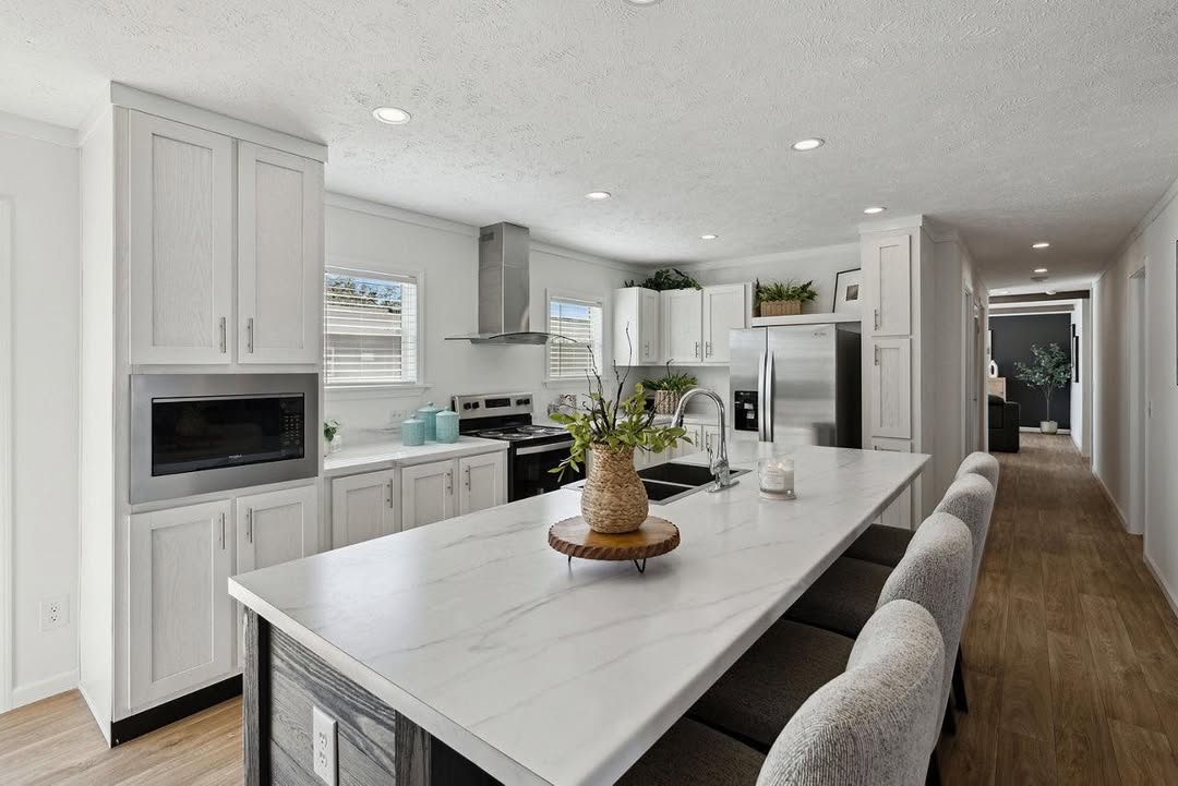 Modern kitchen with white cabinets, marble island counter, and gray barstools. Stainless steel appliances and plants add a fresh touch. Bright, airy ambiance.