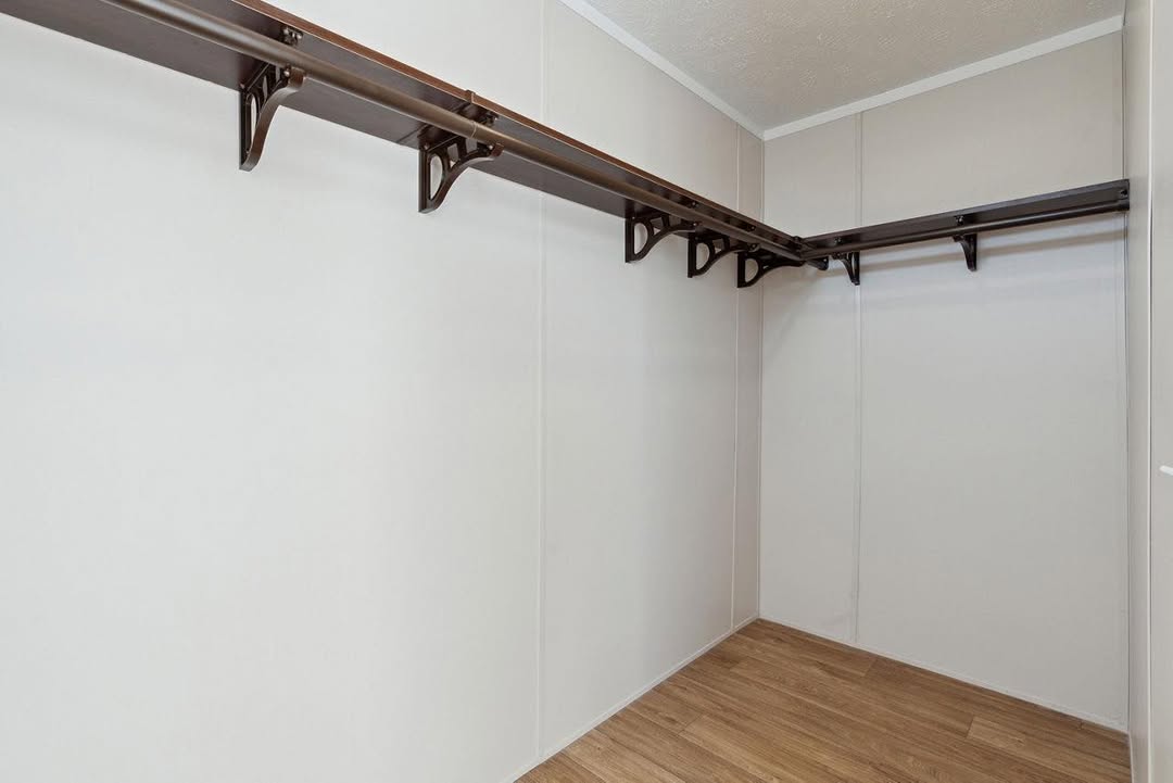 An empty walk-in closet with light beige walls and wooden floor. It features a long, dark wooden shelf with brackets on two sides at head height. Bright and minimal.