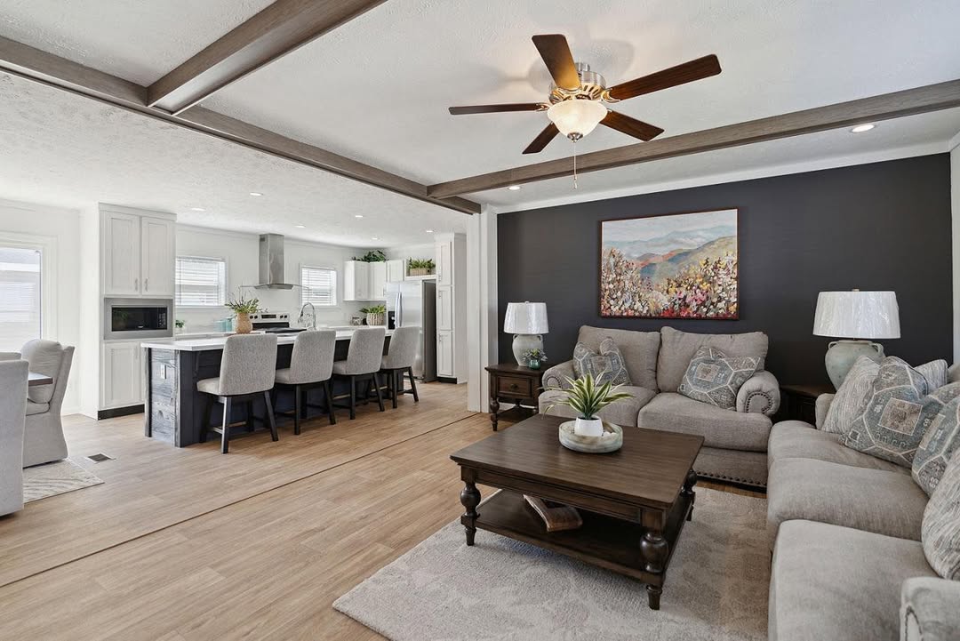 Spacious living room and kitchen with wooden beams. Cozy sofas face a dark accent wall with artwork. Dining area has elegant chairs. Warm, inviting tone.