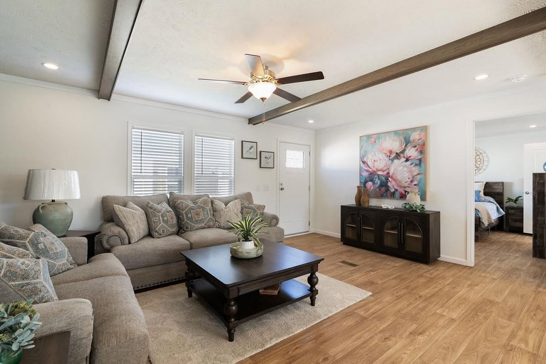 A cozy living room with a beige sectional sofa, decorative cushions, and a dark wood coffee table. A ceiling fan and floral artwork add warmth. A glimpse into a bedroom is visible.