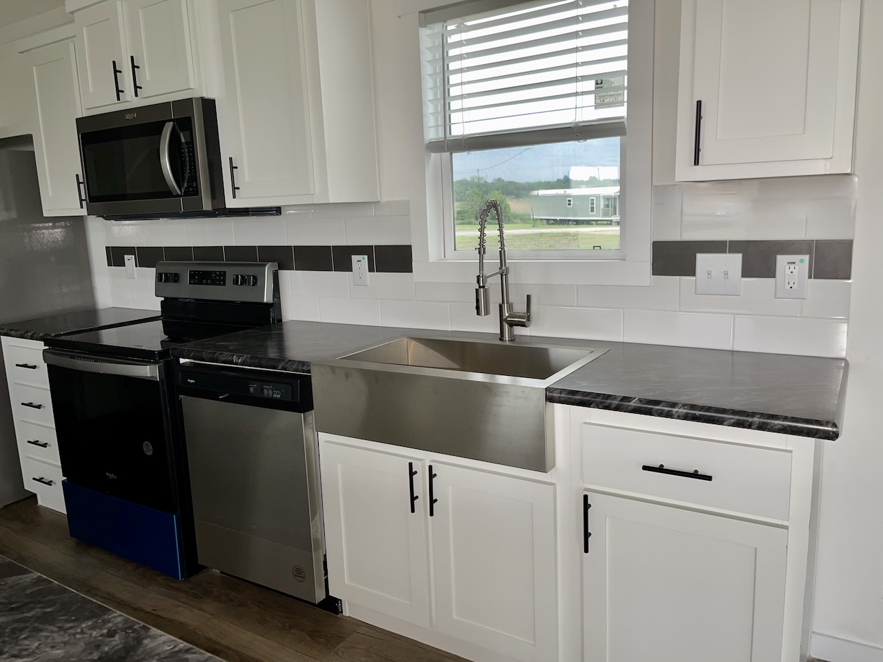 Modern kitchen with white cabinets and black handles, featuring stainless steel appliances, a farmhouse sink, and a window with blinds.