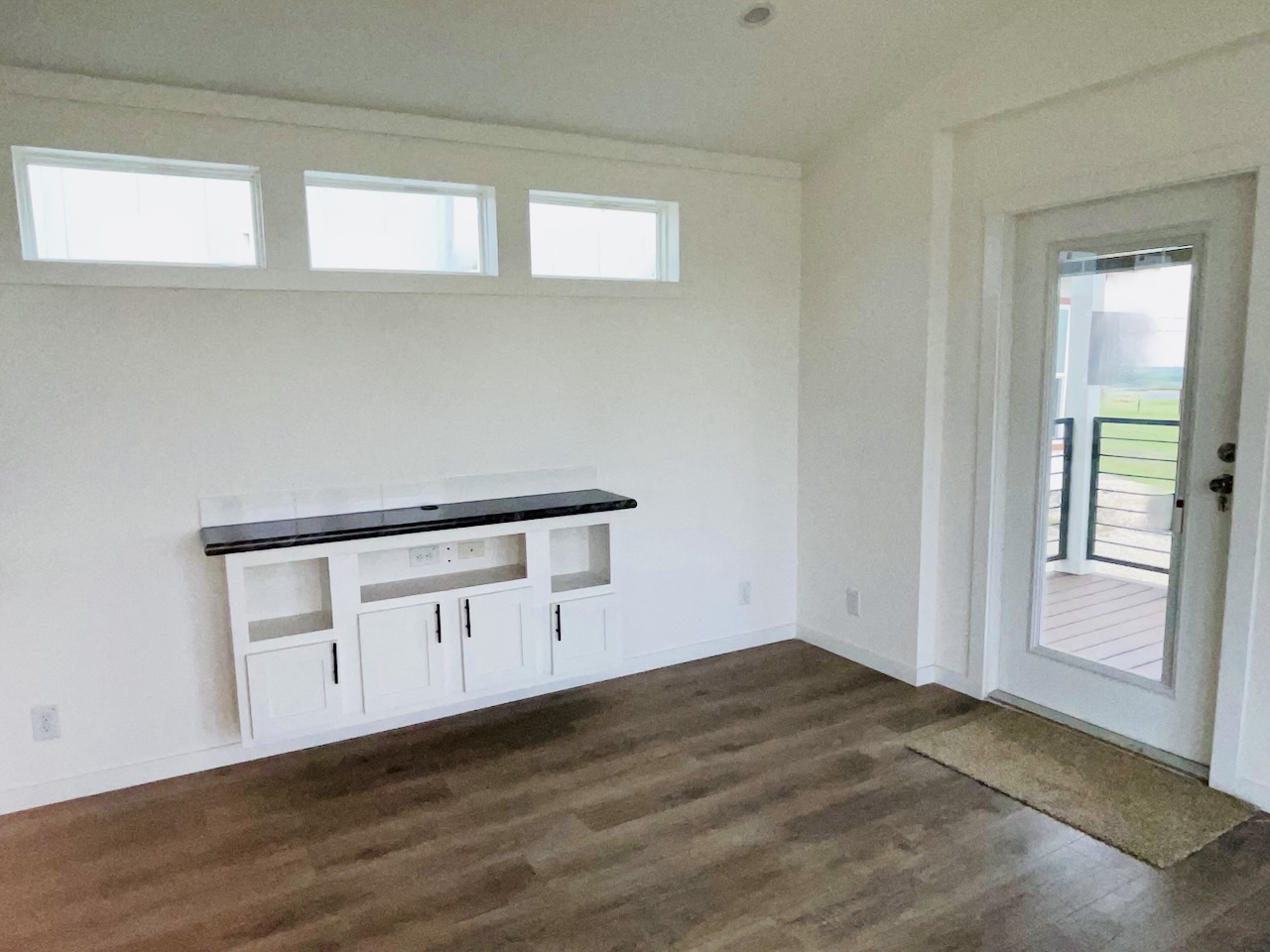A bright, empty room features light wood flooring, white walls, and three small horizontal windows. A built-in white cabinet with a dark countertop is on the left. A glass door on the right leads outside, adding an inviting feel.