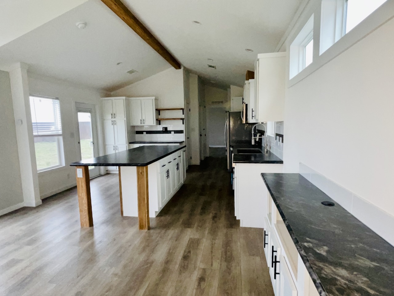 Spacious kitchen with white cabinets, dark countertops, and wood flooring. Bright lighting from large windows and a vaulted ceiling with a wood beam creates an airy atmosphere.