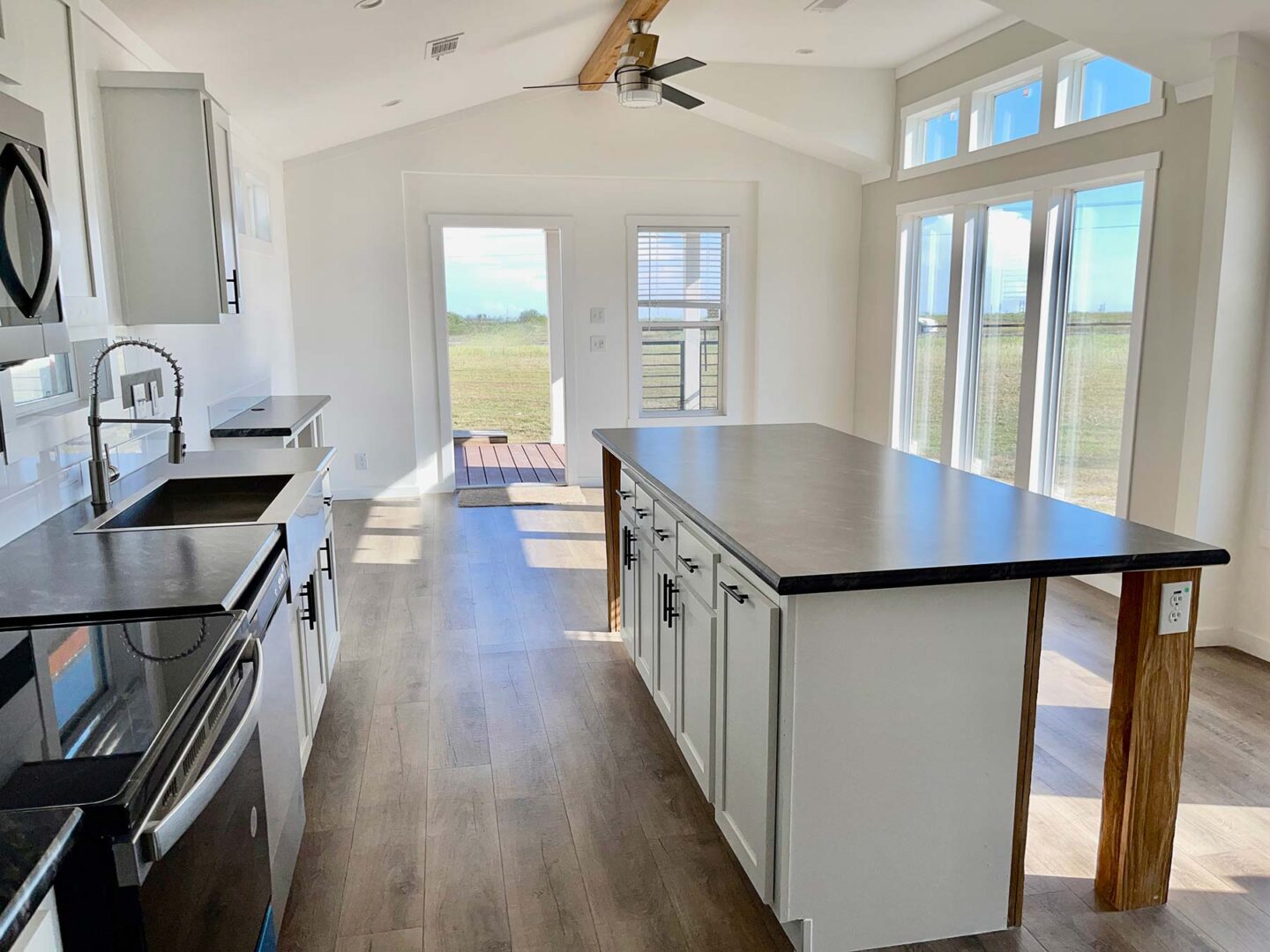 Bright, modern kitchen with a large island featuring white cabinets and dark countertops. Sunlight streams in through expansive windows, offering a serene view of a grassy field.