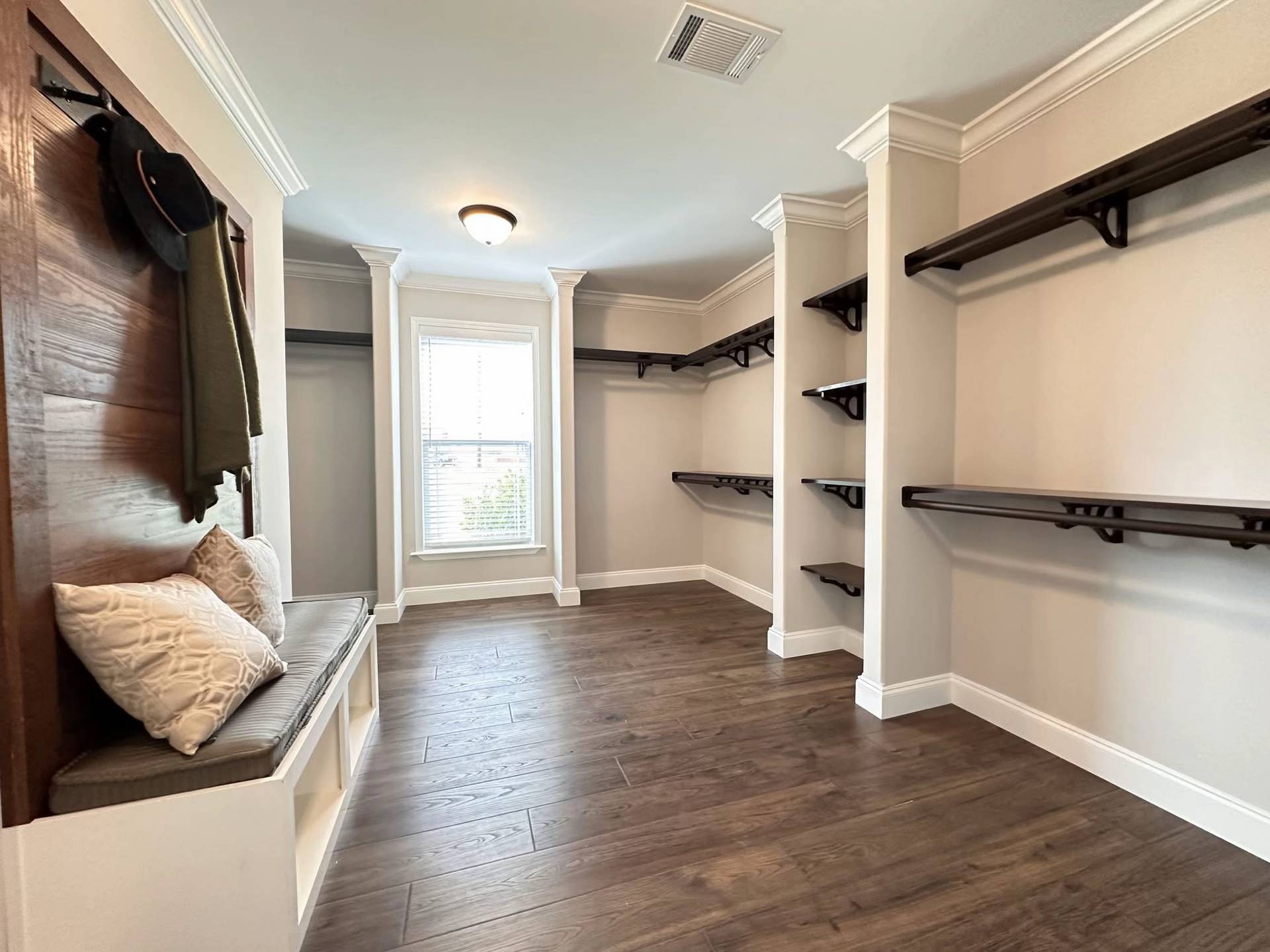 Spacious walk-in closet with dark wooden shelves, light gray walls, and hardwood floors. A window with blinds lets in natural light, creating an airy atmosphere.