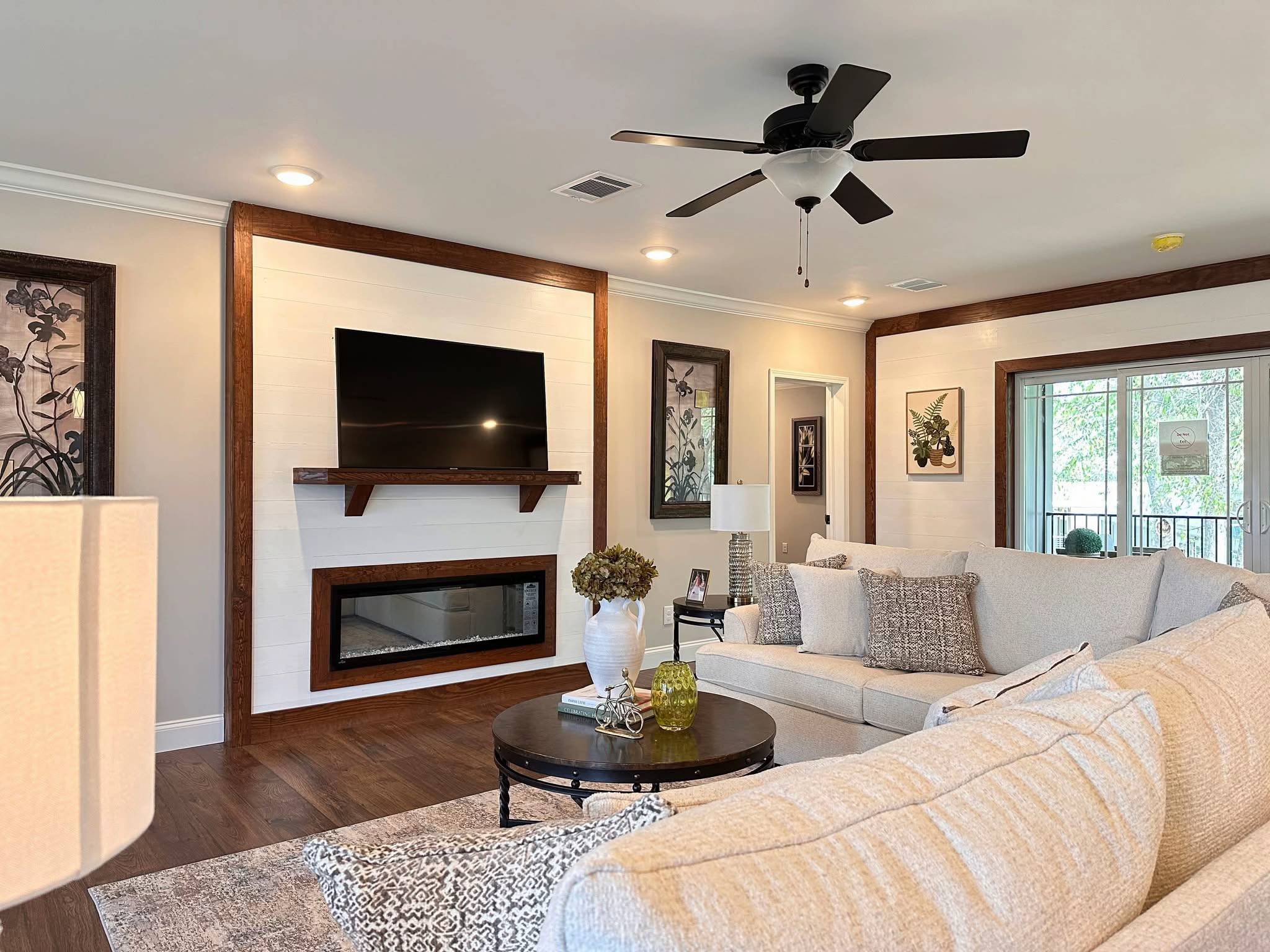 Stylish living room with beige sectional, dark wood accents, wall-mounted TV, and fireplace. Cozy decor, soft lighting, ceiling fan overhead.