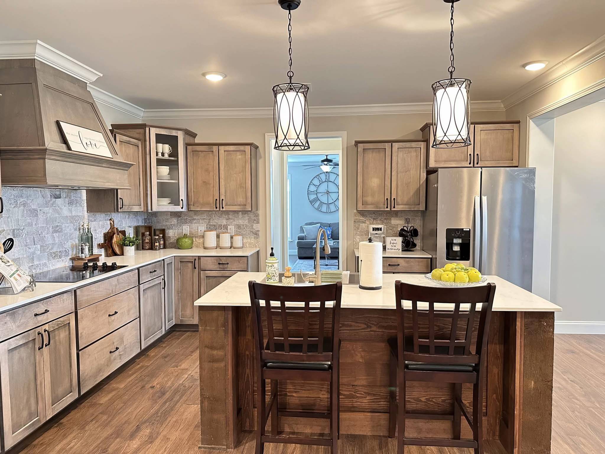 Warm, inviting kitchen with wooden cabinets, island with two chairs, modern lighting, stainless steel fridge, and accents of greenery and fruit.