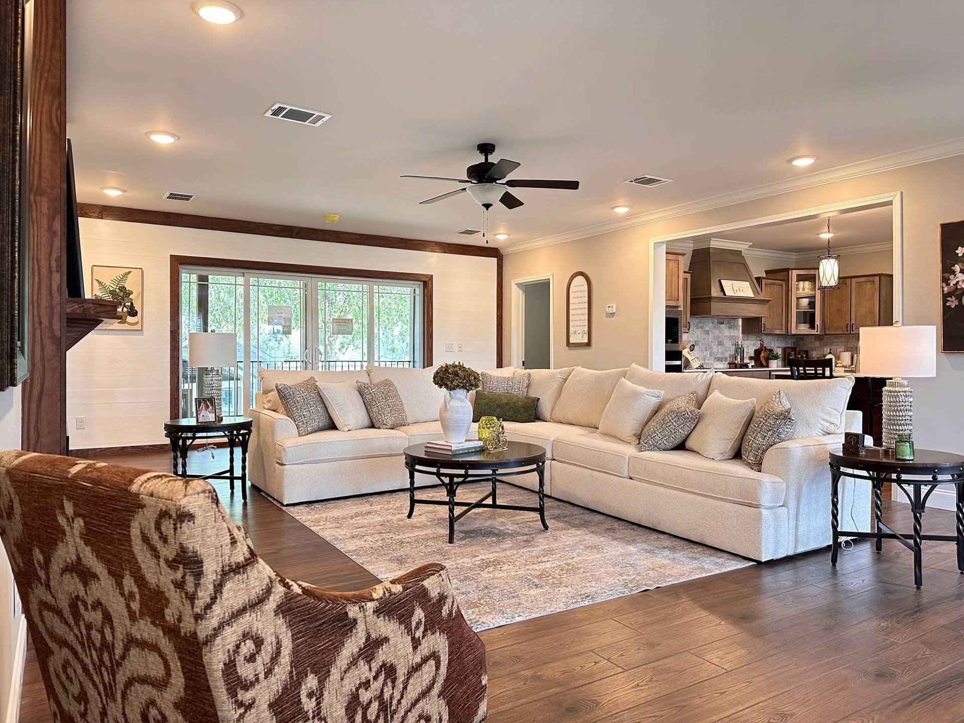 Spacious living room with a beige sectional sofa, patterned armchair, coffee tables, and a rug on wooden floor. Warm lighting and a cozy atmosphere.