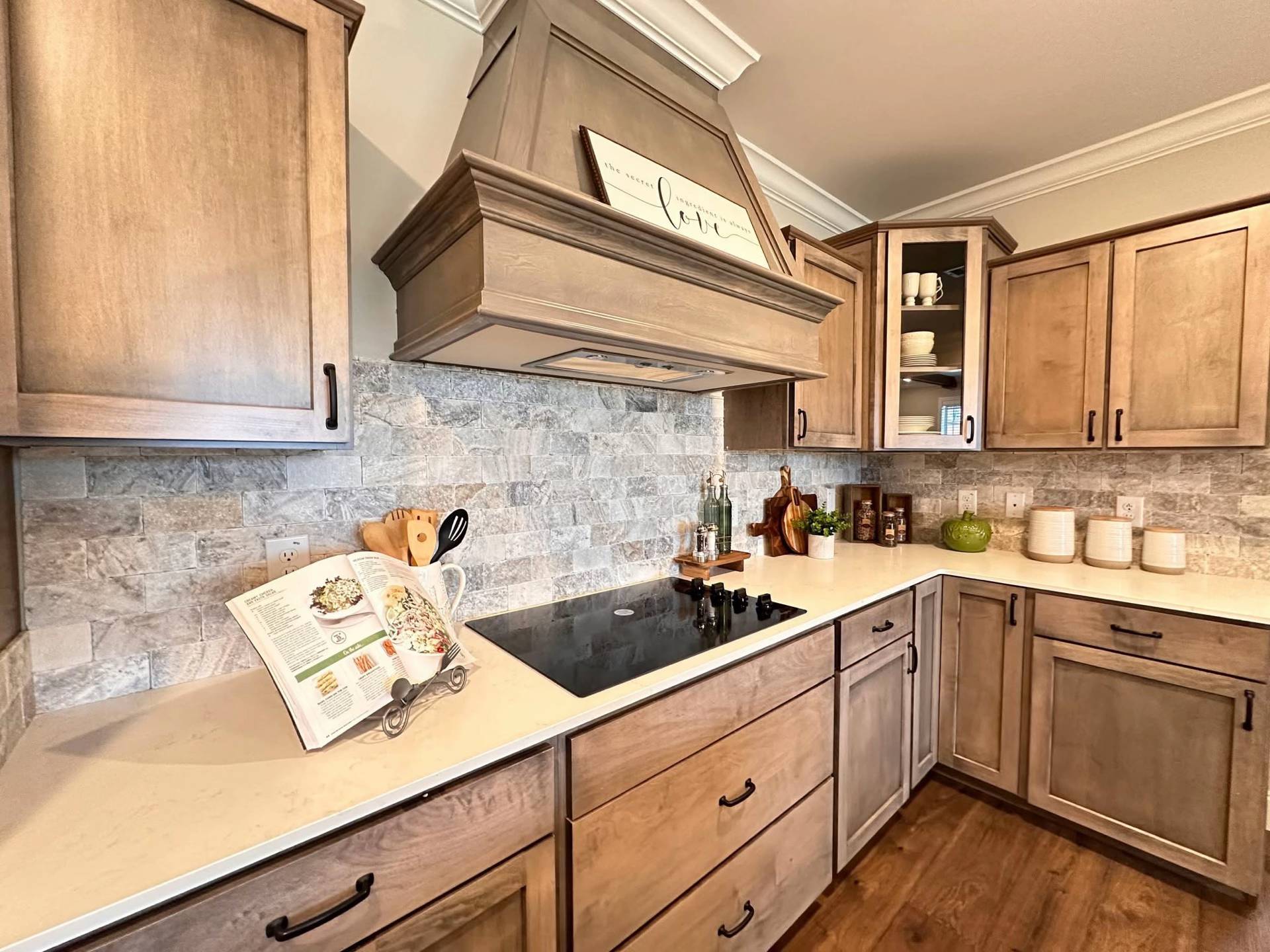 A cozy kitchen with wooden cabinets and stone backsplash. An open cookbook rests on the counter by a modern stovetop. The mood is warm and inviting.