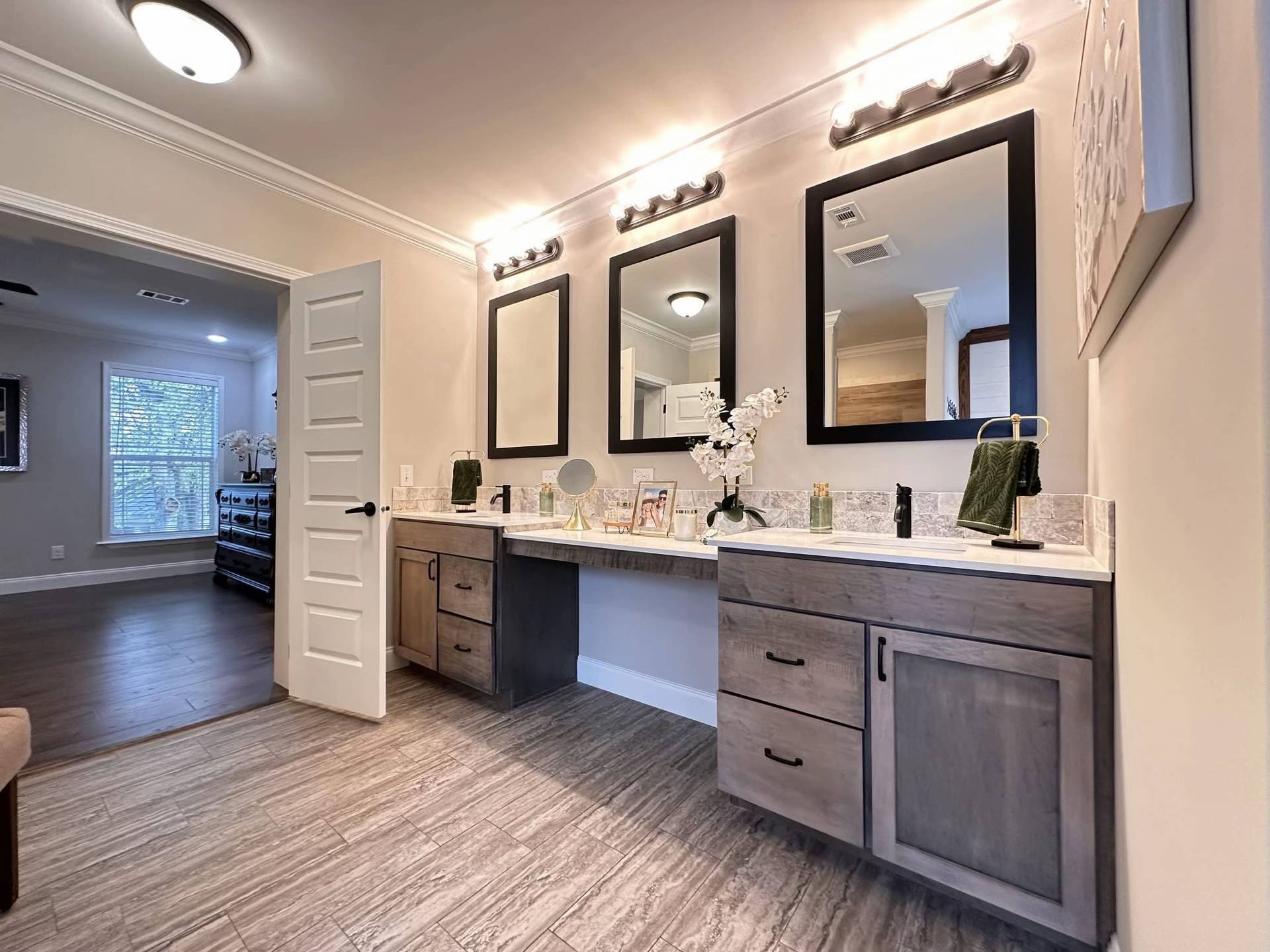 Spacious bathroom with twin gray vanities, large mirrors, and bright lights. Warm wood flooring leads to an open door revealing a sunlit room. Cozy and elegant.