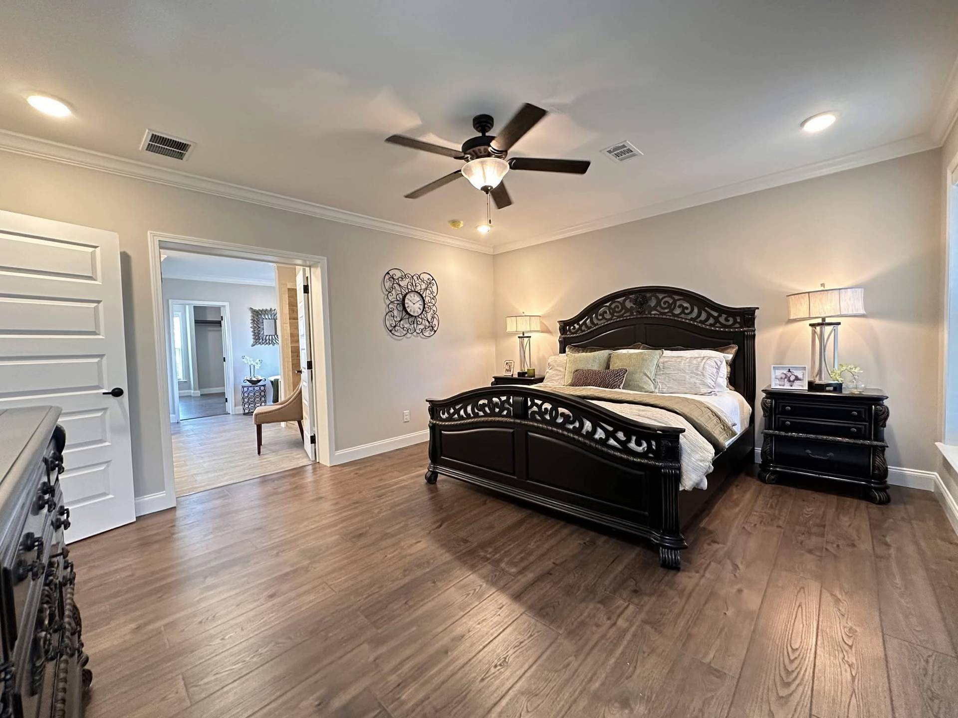 Spacious bedroom with dark wood furniture, including an ornate bed and nightstands. Soft lighting, ceiling fan, and open doorway to a bright hallway.