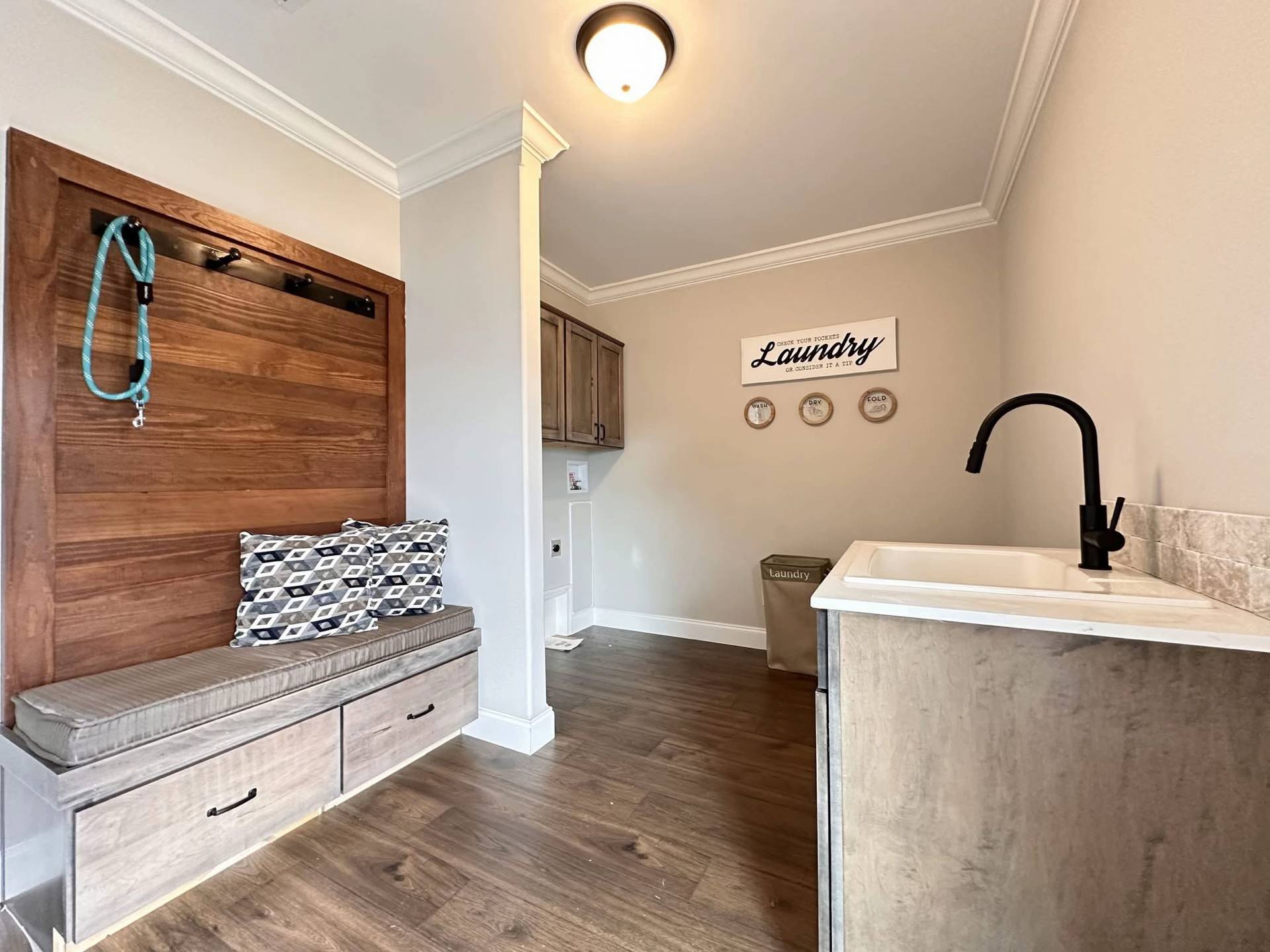 Bright laundry room with wood floors, a bench with cushions, and a hanging leash. Overhead light, wooden cabinetry, sink with black faucet, and "Laundry" sign. Cozy and organized.