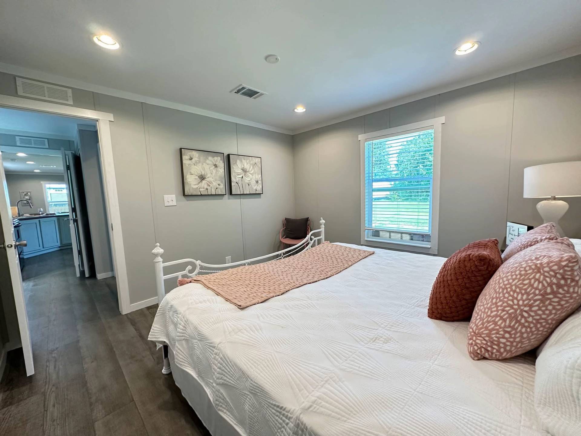 A cozy bedroom with a white bed, pink cushions, and a soft blanket. There's soft lighting, floral wall art, a window view, and a serene ambiance.