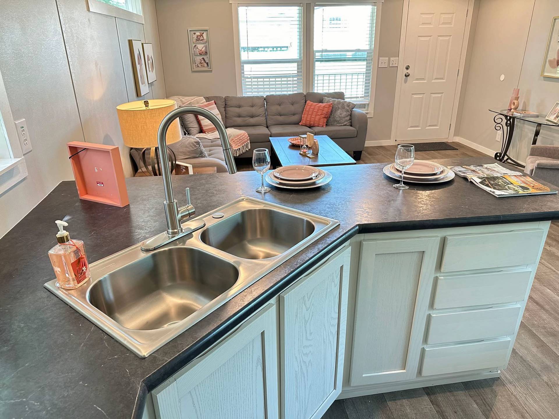 Modern interior with a kitchen island featuring a double sink and black countertop. In the background, a cozy living area with a gray sofa, orange accents, and set dining table near large windows.