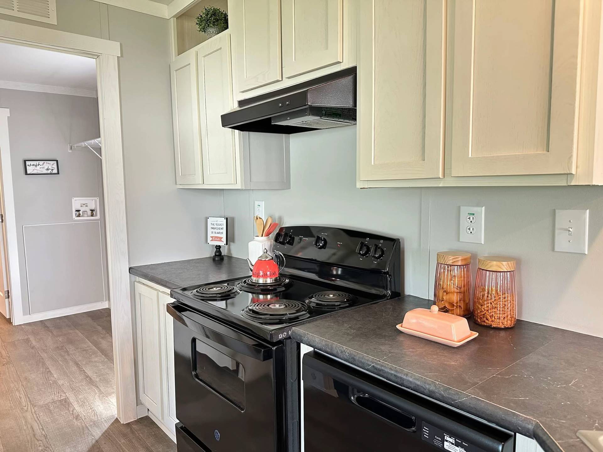 A modern kitchen with black appliances, light cabinetry, and dark countertops. A red kettle sits on the stovetop, and jars of cookies line the counter.