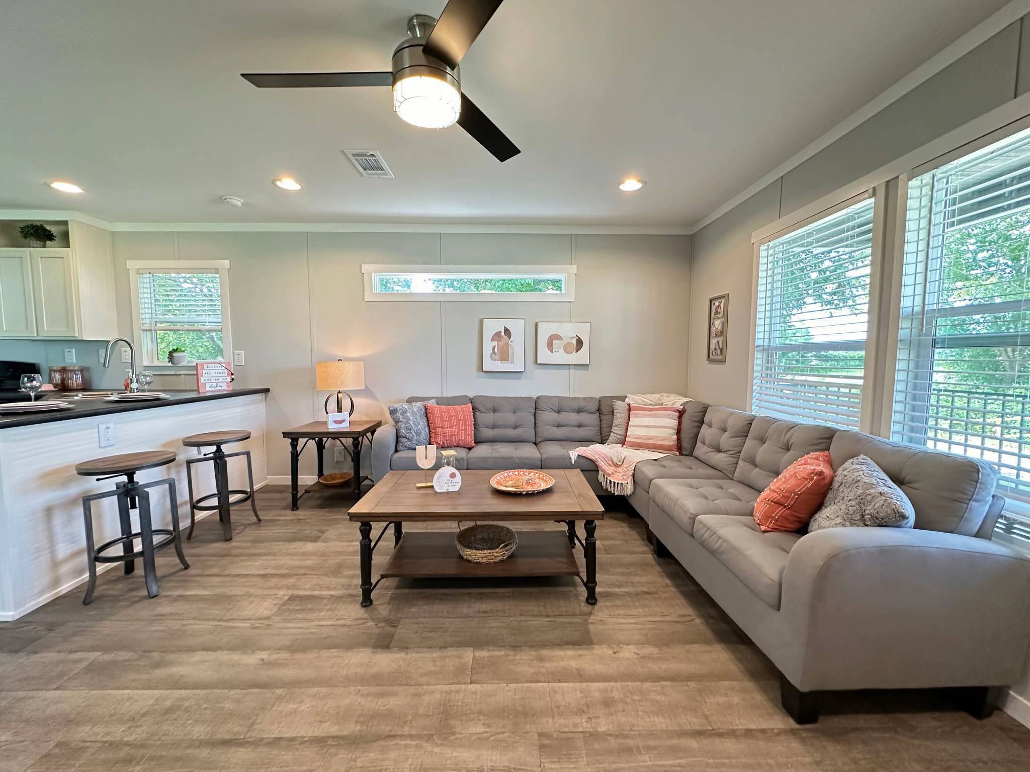 Modern living room with gray sectional sofa adorned with pink and gray pillows. Wooden coffee table, bar stools, and light wood flooring add warmth.
