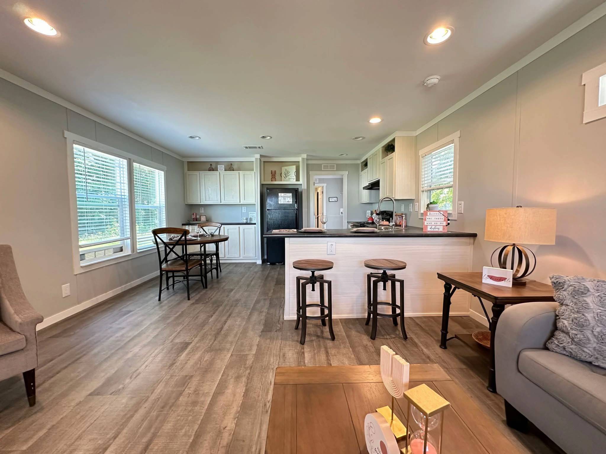 Modern open-plan living room and kitchen with light gray walls and wood flooring. Features a kitchen island, barstools, dining table, and cozy seating.