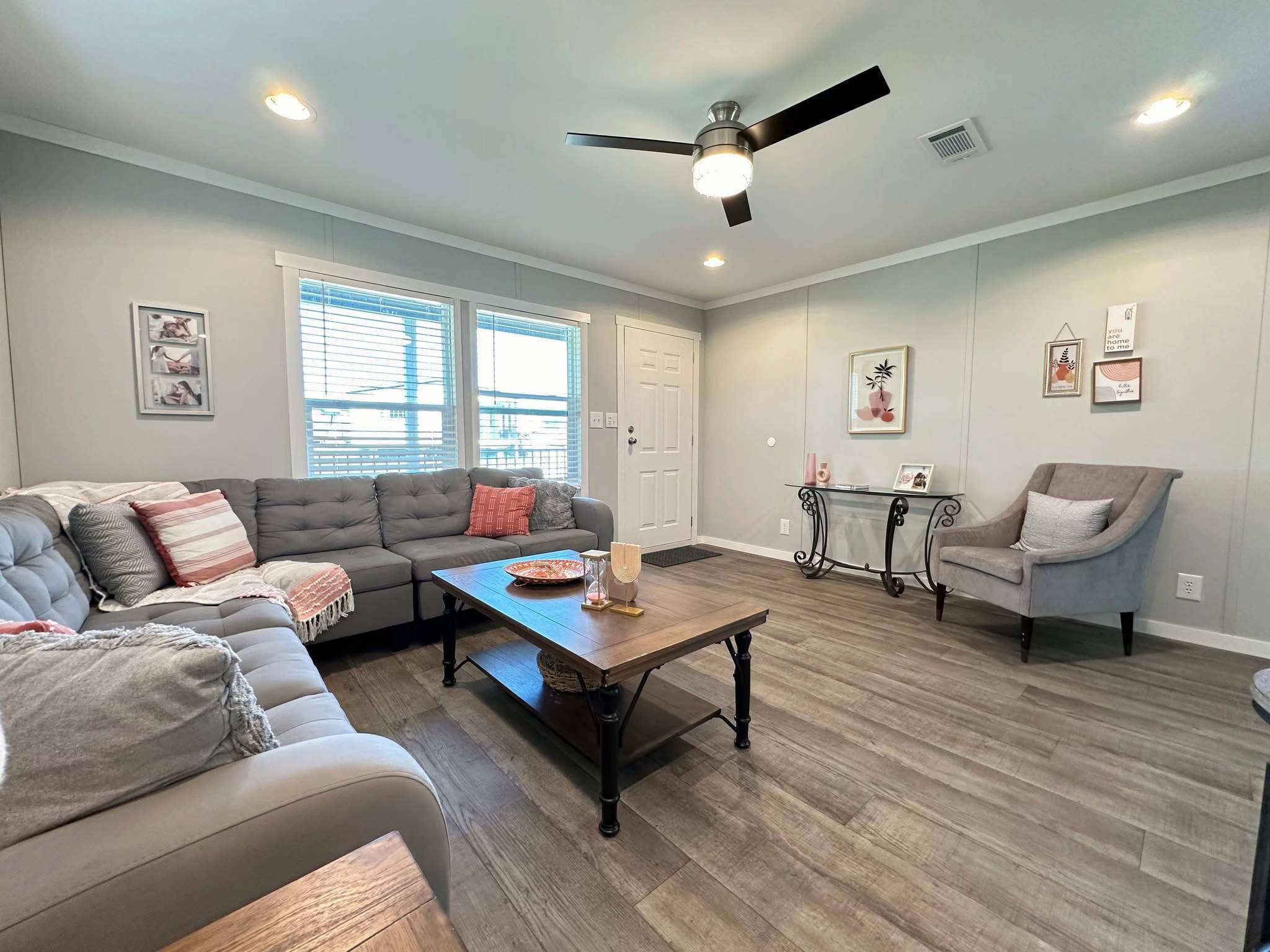 Cozy living room with gray sofas, colorful cushions, a brown coffee table, and a plush armchair. Soft lighting creates a warm, inviting atmosphere.