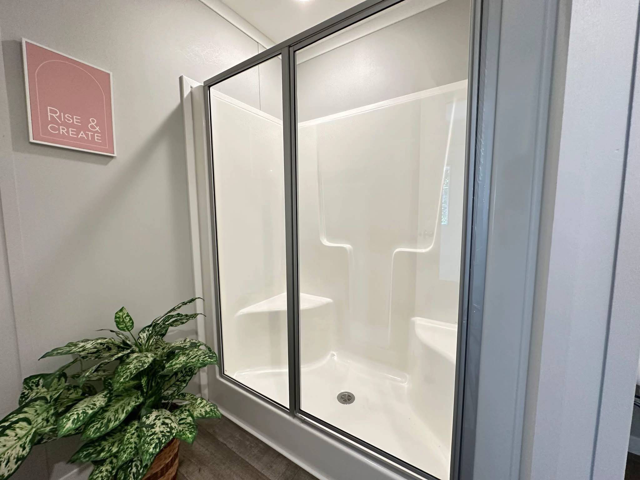A clean, modern shower with glass doors and built-in white seats is next to a plant. A "Rise & Create" sign hangs on the light gray wall.