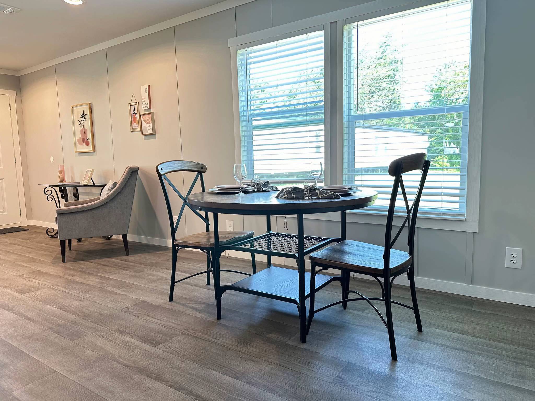 Modern dining area with a round wooden table set for two. Metal chairs are placed beside it, near a window with blinds, creating an inviting ambiance.