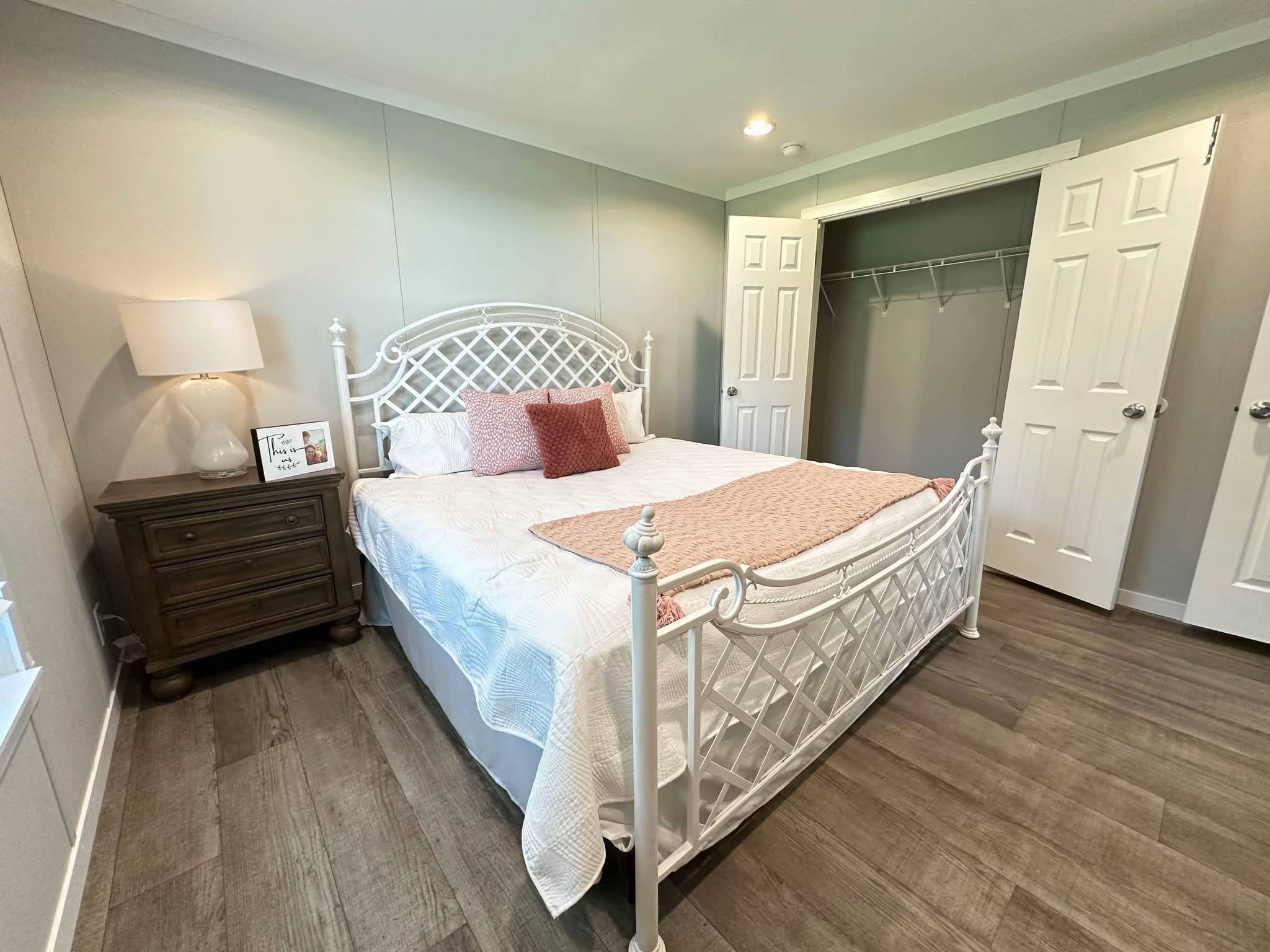A cozy bedroom with a white metal bedframe, white bedding, and pink accent pillows. A wooden nightstand with a lamp is on the left. Open closet doors reveal an empty closet, while soft lighting creates a warm ambiance.