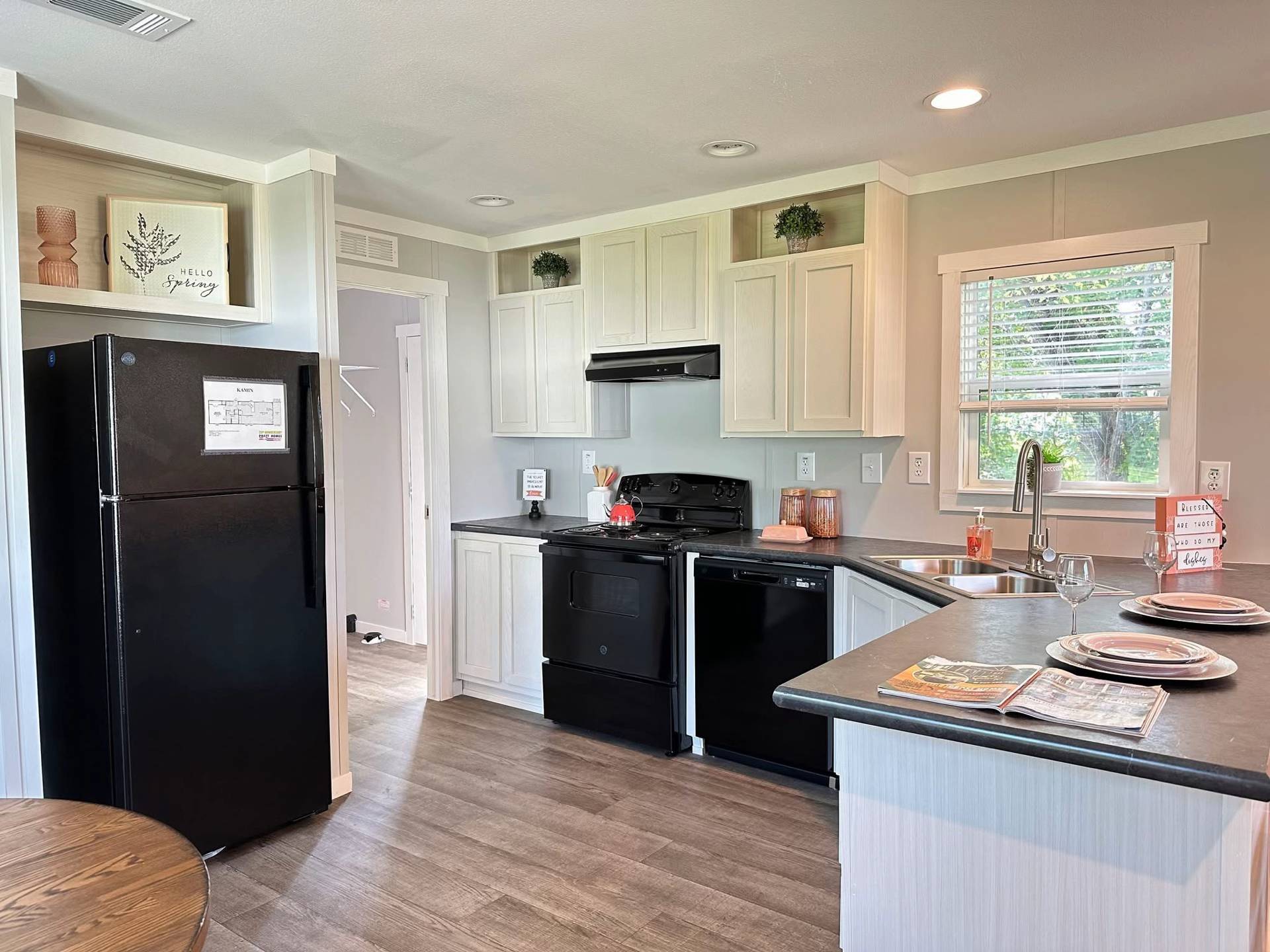 A modern kitchen with light cabinetry, black appliances, and wood floors. A window provides natural light, and a small dining area is set with plates.