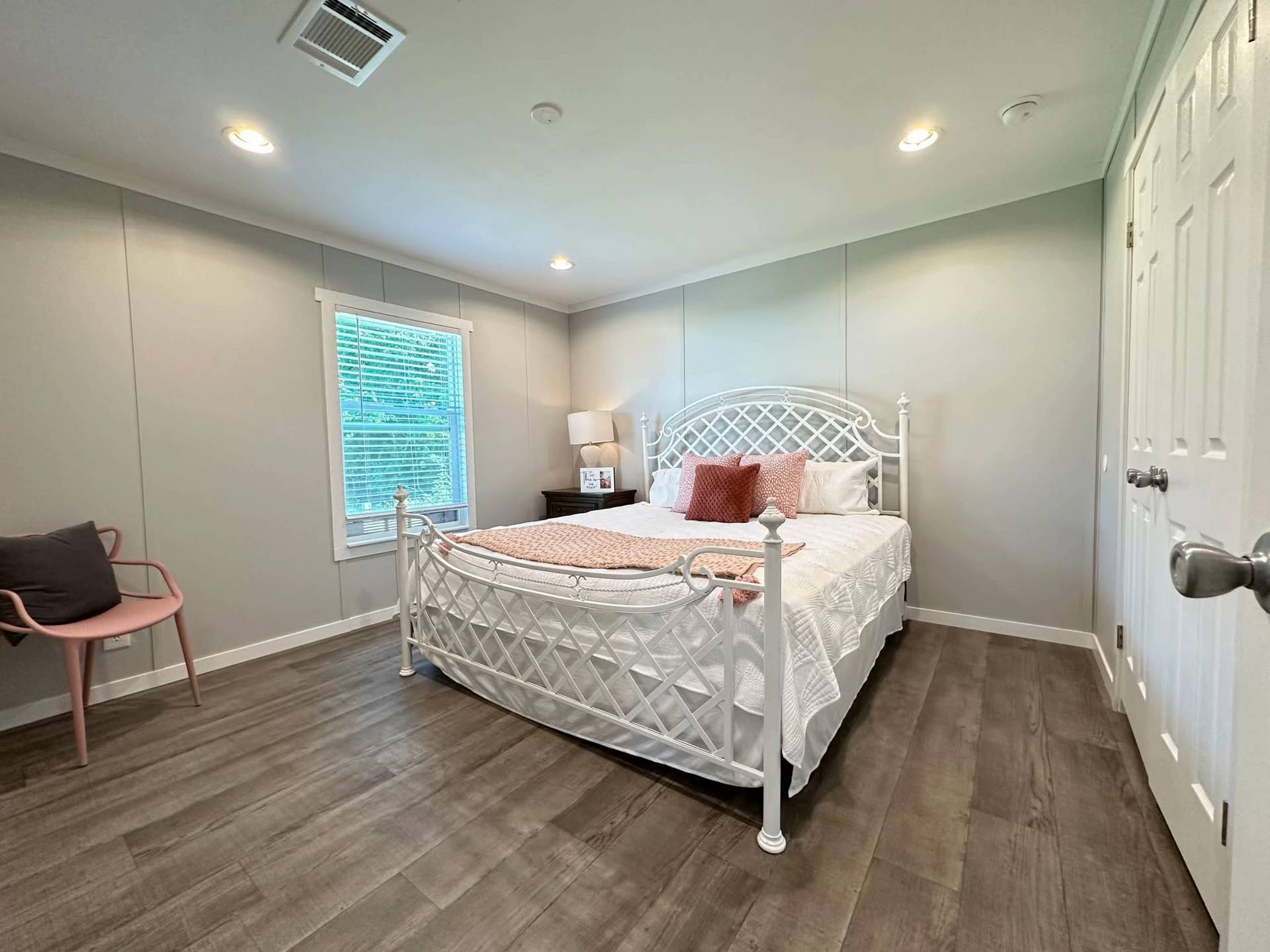 Bright bedroom with a white metal bed, pink and white bedding, wooden floor, and soft lighting. A window with blinds, a pink chair, and a nightstand with a lamp add cozy touches.