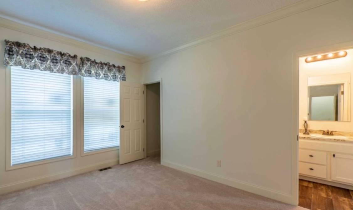 A bright, empty room with beige carpet and cream walls. Two large windows with patterned valances let in natural light. An open door leads to a lit bathroom.