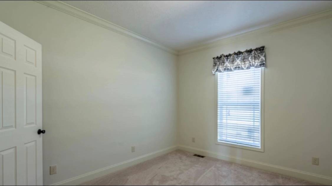 A small, empty room with white walls and beige carpet. A window with a patterned valance lets in soft light, creating a calm, inviting atmosphere.