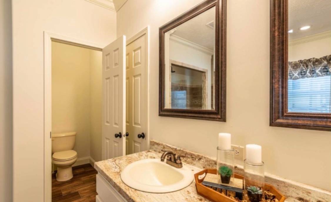 Bright bathroom with a granite countertop, a white sink with bronze faucet, and a large framed mirror. Decorative plants and candles sit on a wooden tray, with a doorway leading to a toilet and wood flooring visible in the background.