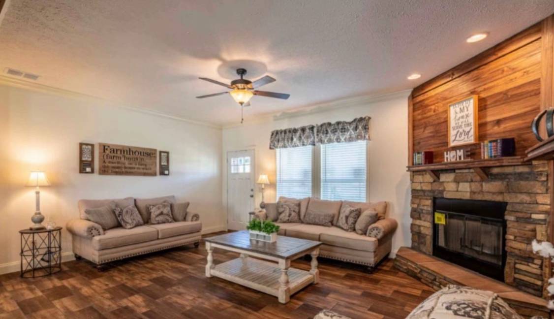 A cozy living room with two beige sofas, a wooden coffee table, and a stone fireplace. A ceiling fan is above, and a sign reads "Farmhouse" on the wall.