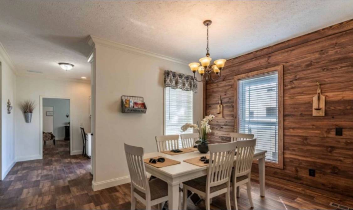A cozy dining room with a wooden table and four chairs, adorned with a potted plant. The room features a wood-paneled accent wall, a window with blinds, and a chandelier overhead, creating a warm, inviting atmosphere.
