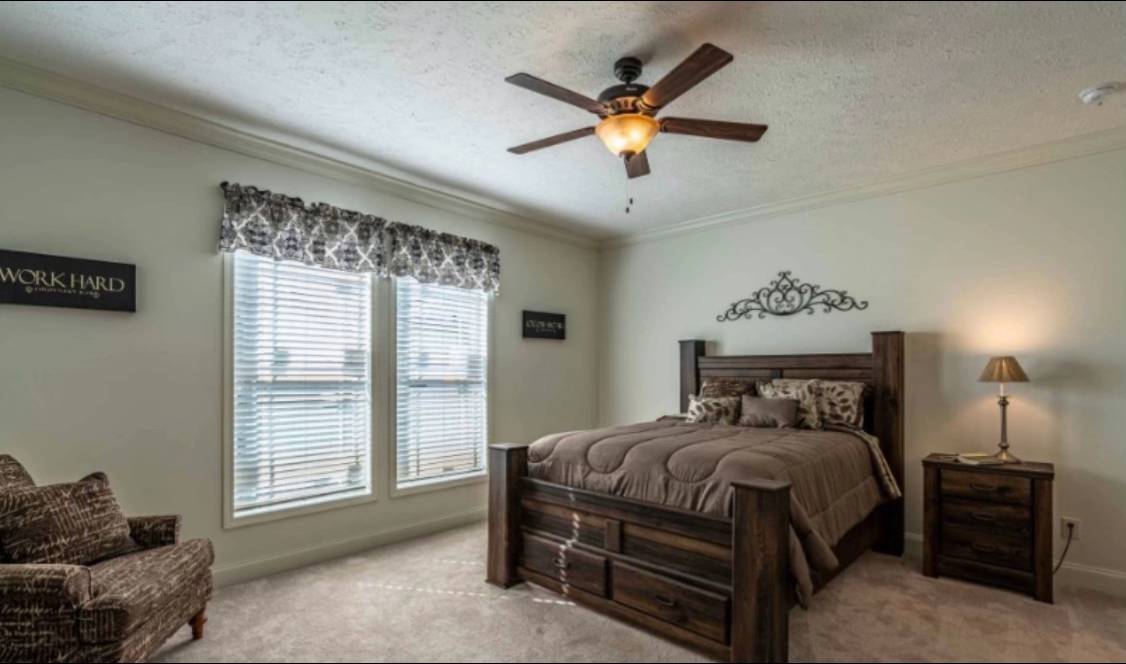 A cozy bedroom with a wooden bed, brown bedding, and matching side table. A ceiling fan hangs above, natural light streams through two windows, creating a warm and inviting atmosphere. Decorative signs and a patterned chair enhance the decor.
