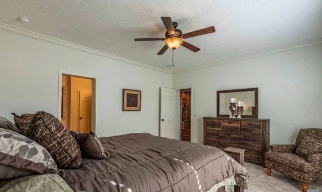 A cozy bedroom with a large bed featuring patterned brown pillows, a wooden dresser with a mirror, an armchair, and a ceiling fan casting a warm light.